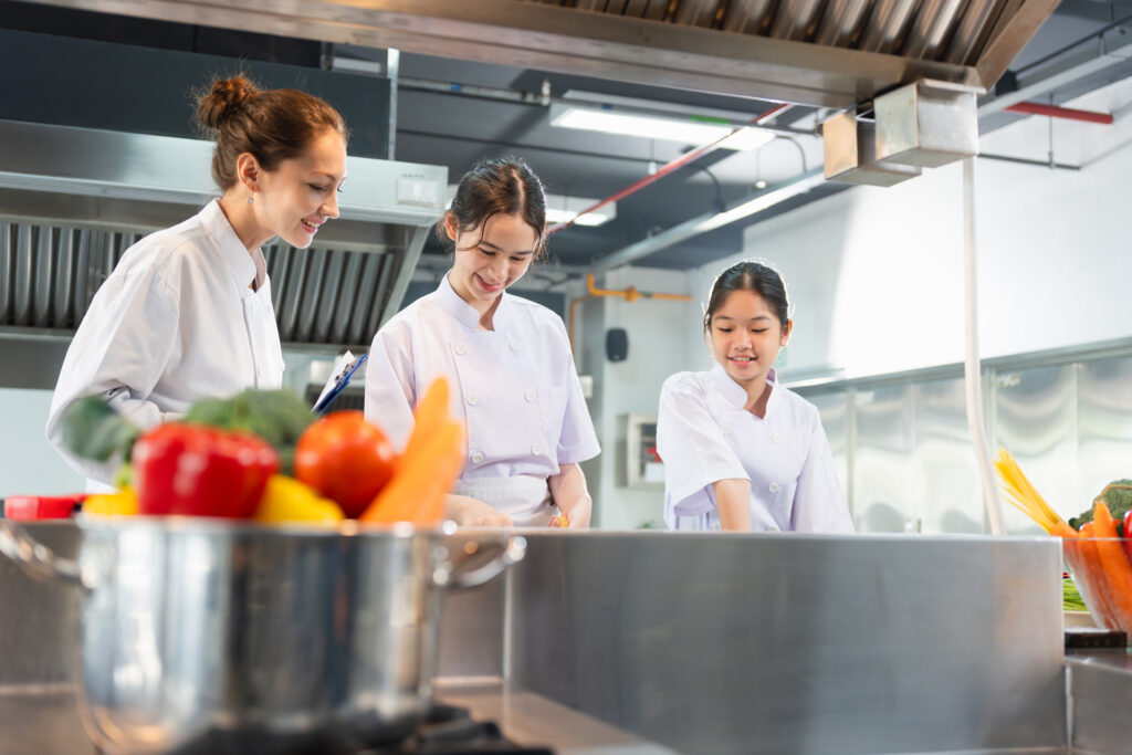 culinary-instructor-guides-students-professional-kitchen-young-chefs-learning-cook-with-fresh-ingredients-women-chef-uniforms-preparing-food-commercial-kitchen